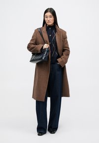 Cappotto di lana marrone con grandi bottoni indossato sopra un maglione blu navy con un motivo a rombi. Abbinato a jeans a gamba larga e una borsa blu navy.