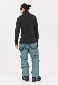 Langärmliges schwarzes Oberteil und türkisfarbene Schneehosen. Die Hosen haben kontrastierende schwarze seitliche Akzente und verstellbare Träger. Schwarze Schneestiefel sind sichtbar.