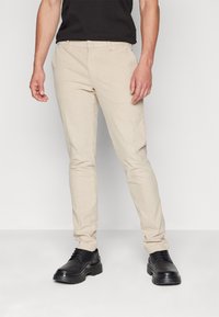 Béžové úzké kalhoty s texturovanou látkou a předními kapsami, doplněné černými koženými šněrovacími botami s tlustou podrážkou.