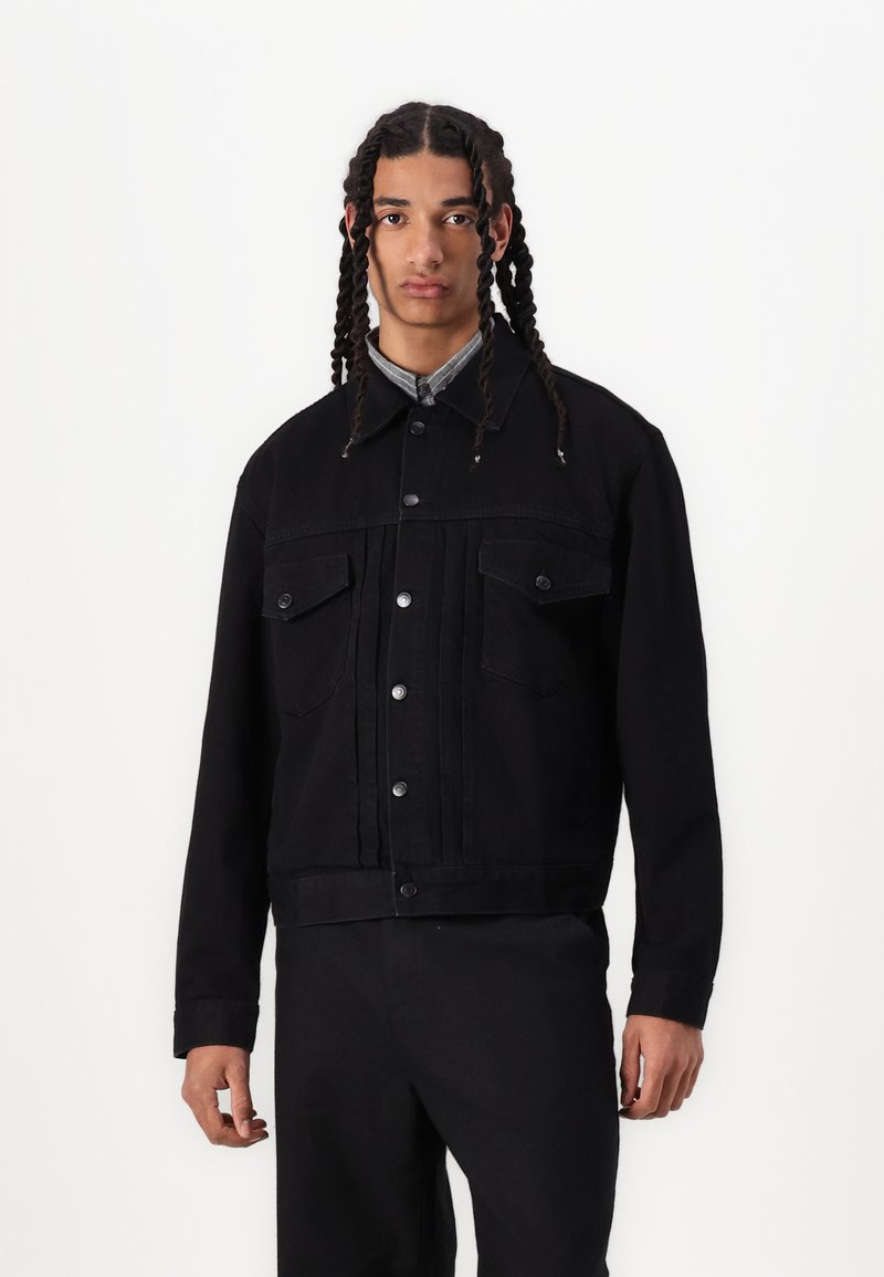 Svart denimjakke med kragedesign, frontlommer med klaff og metallknapper. Tøyet har en glatt tekstur og strukturert passform.