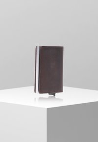 Portefeuille en cuir marron avec un design élégant et minimaliste, agrémenté d'accents en argent. Présente des coutures le long des bords et une forme rectangulaire compacte.