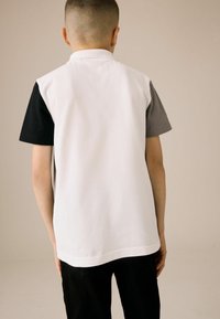 Polo avec un corps blanc, des manches courtes noires et des accents gris. Fabriqué en tissu lisse, avec un col classique et une ourlet droit.