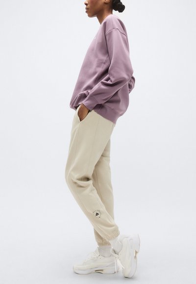 Μωβ φούτερ με άνετη γραμμή, συνδυασμένο με μπεζ τζόγκερ με πλαϊνές τσέπες και λογότυπο στο αριστερό πόδι. Το σύνολο ολοκληρώνουν λευκά αθλητικά παπούτσια.