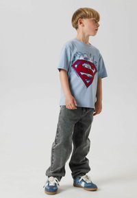 Chłopiec stoi bokiem, ubrany w jasnoniebieską koszulkę Superman, szare dżinsy i niebieskie trampki. Neutralne tło podkreśla jego strój.