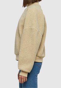 Beige Fleece-Pullover mit hohem Kragen, überschnittenen Schultern und gerippten Bündchen. Weiche Textur und lässige Passform, kombiniert mit blauen Jeans.