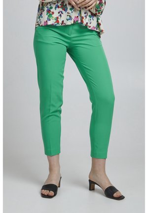 Pantalons ajustés verts en tissu lisse, avec un design tapered. Associés à des escarpins noirs ouverts pour un accent de contraste.