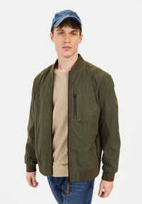 Olivengrøn bomberjakke med en lynlåslomme på brystet, rib-strikkede manchetter og kant, båret over en beige sweatshirt og parret med en blå kasket.