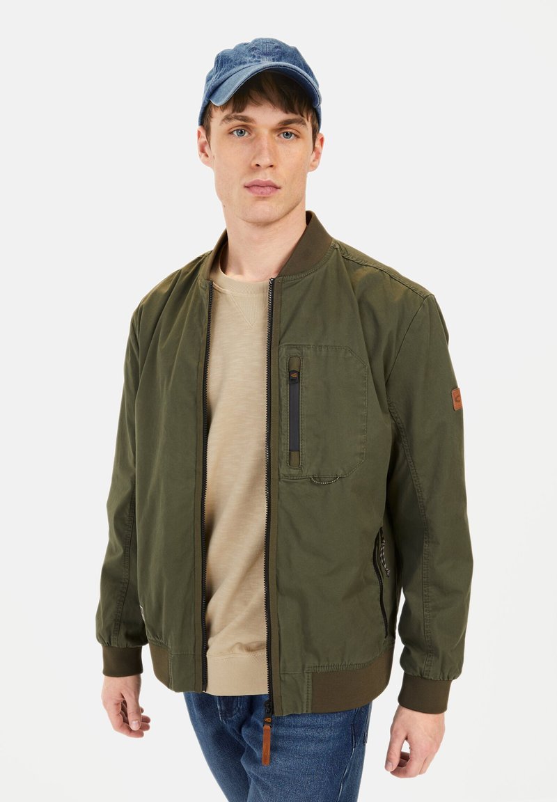 Olivengrøn bomberjakke med en lynlåslomme på brystet, rib-strikkede manchetter og kant, båret over en beige sweatshirt og parret med en blå kasket.