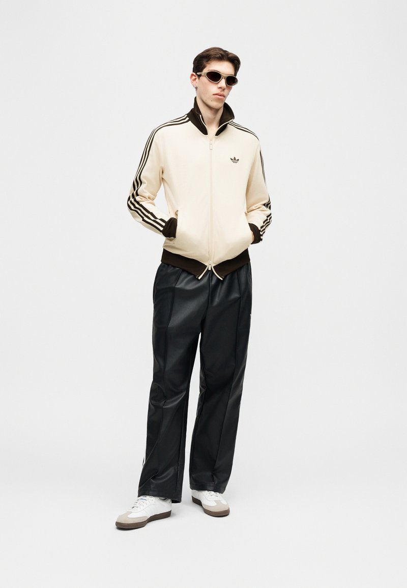 Hombre joven con chaqueta deportiva crema y marrón, pantalones de cuero negros, zapatillas blancas y gafas de sol, de pie con las manos en los bolsillos.