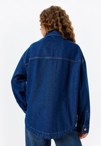 Chaqueta de mezclilla de color azul oscuro con un corte relajado, que presenta un collar puntiagudo, mangas largas y detalles cosidos en el dobladillo y los puños.