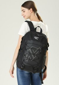 Zaino nero con superficie testurizzata, caratterizzato da un design con cordino elastico e logo bianco. Include spallacci regolabili e maniglia superiore.