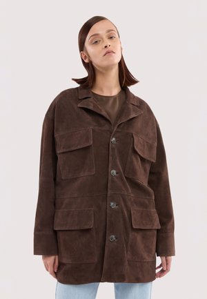 Frau trägt eine übergroße dunkelbraune Wildlederjacke mit Frontknöpfen und vier großen Klappentaschen, kombiniert mit hellblauen Jeans.