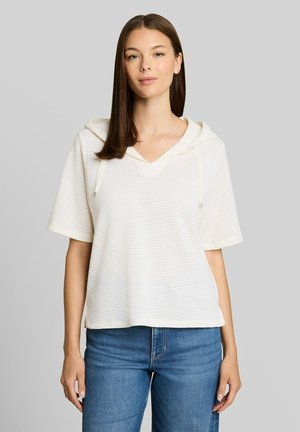 Jonge vrouw met stijl bruin haar die een witte, gestructureerde korte mouwen hoodie en blauwe spijkerbroek draagt, staand tegen een effen grijze achtergrond.