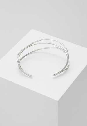 Silbernes offenes Manschettenarmband mit drei dünnen parallelen Bändern, das auf einem weißen geometrischen Sockel vor grauem Hintergrund liegt.