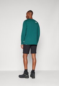 Chemise à manches longues turquoise, shorts noirs et bottes de randonnée noires. Présence d'un logo dans le haut du dos. Matériau doux et texturé. Coupe décontractée.