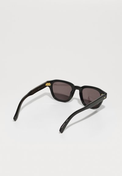 Black sunglasses with a square frame, dark lenses, glossy finish, and gold accent on the temples. Slim, curved black arms extend from the frame.