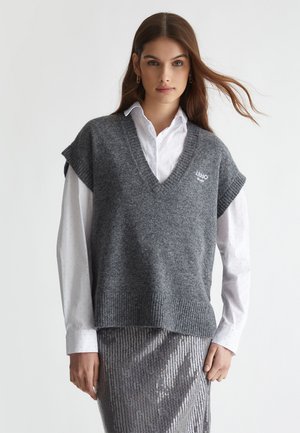 Maglione canotta grigio a maglia con scollo a V e maniche corte, con orlo a costine e dettaglio del logo, indossato sopra una camicia bianca con colletto.
