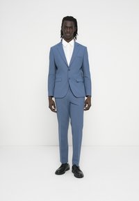 Ανοιχτό μπλε κοστούμι με μονόπιετο σχέδιο, πέτο με εγκοπές και πλαϊνές τσέπες. Συνδυασμένο με λευκό πουκάμισο και μαύρα παπούτσια. Λεία υφή.