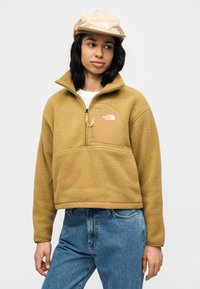 Νεαρός που φοράει μια μπεζ φλις μπλούζα με φερμουάρ στην τσέπη στο στήθος και αντίστοιχο καπέλο, σε συνδυασμό με μπλε τζιν, μπροστά σε απλό φόντο.