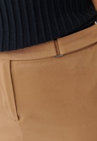 Pantalon beige avec une texture lisse, une coupe ajustée et un accent subtil à la taille. Associé à un pull en maille bleu marine.