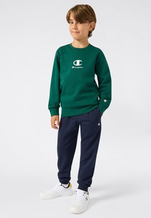 Sweatshirt vert avec logo blanc, associé à un pantalon de survêtement bleu marine et des baskets blanches avec des accents verts et jaunes. Tenue de sport décontractée.
