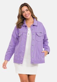 Lila Jeansjacke mit zwei Brusttaschen, silbernen Beschlägen und weißen Nähten. Über einem weißen Kleid getragen, das eine lockere Passform und einen Knopfverschluss hat.