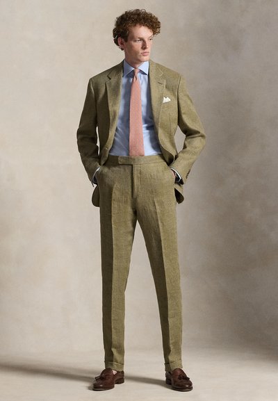 Mann trägt einen olivgrünen Anzug mit hellblauem Hemd, rosa gemustertem Krawatte, weißem Einstecktuch und braunen Tassel-Loafers, steht mit den Händen in den Taschen.