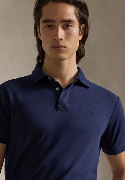 Junger Mann mit schulterlangem dunklem Haar trägt ein marineblaues Poloshirt mit einem kleinen gestickten Logo auf der Brust vor neutralem Hintergrund.