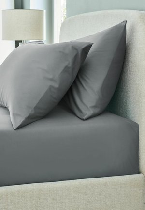 Two dark gray pillows on a matching fitted sheet with a light gray upholstered bed frame and a white lamp in the background.