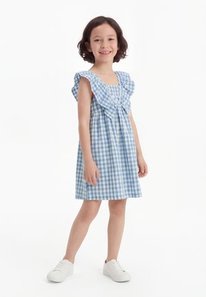 Niña sonriente vestida con un vestido azul y blanco de cuadros con mangas con volantes y zapatillas blancas, de pie sobre un fondo blanco.