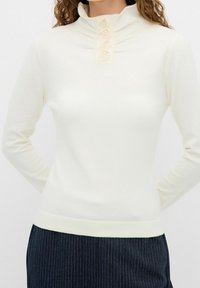 Biała bluzka z długim rękawem i wysokim kołnierzem ozdobionym czterema haftowanymi guzikami w kształcie róż, o gładkiej fakturze i dopasowanym kroju, noszona z ciemnymi spodniami w paski.