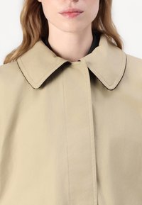 Beżowy płaszcz z dużym, zaokrąglonym kołnierzem, z zapięciem na guziki i kontrastującymi czarnymi przeszyciami. Gładka, lekka faktura materiału.