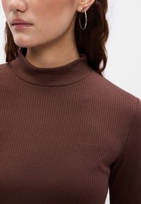 Barna bordázott, magas nyakú top, testhez simuló fazonnal. Jellemzője a kis logó részlet, amely megegyező színű, valamint a sima, textúrált anyag.