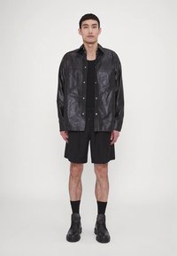 Άντρας που στέκεται φορώντας μαύρο δερμάτινο μπουφάν, μαύρο φανελάκι, μαύρες βερμούδες, μαύρες κάλτσες και μαύρες μπότες με φόντο λευκό τοίχο.