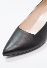 Chaussure plate en cuir noir à bout pointu avec finition texturée et doublure intérieure beige sur fond blanc.