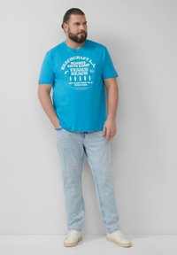 Turquoise T-shirt met korte mouwen en witte grafische print met "Venice Beach" en surfboardontwerpen. Draag het met lichtblauwe jeans en witte sneakers.