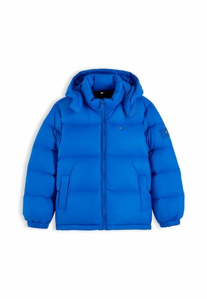 Dreng iført en blå dunjakke, rød sweater, blå jeans og hvide sneakers står med hænderne i lommerne på en hvid baggrund.