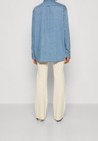 Donna che indossa una camicia di denim azzurro chiaro e pantaloni a gamba larga crema con motivo, in piedi di spalle su uno sfondo grigio uniforme.