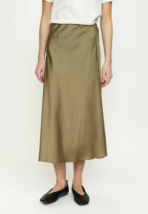 Osoba oblečená v olivovo zelenej midi sukni, bielom tričku s krátkym rukávom a čiernych plochých topánkach, stojaca pred jednoduchým svetlým pozadím.