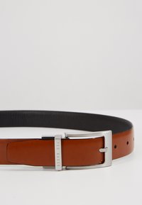 Brun skinnbelte med svart innvendig, rektangulær sølvspenne og preget "TED BAKER"-logo. Har flere justeringshull.