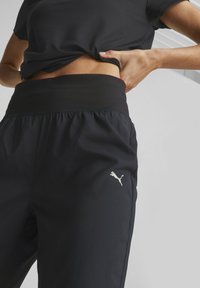 Svarta träningsbyxor med elastisk midja, en subtil glans och en kontrasterande guld Puma-logotyp på vänster sida. Slät textur.