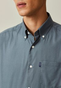 Chemise boutonnée en tissu gris ardoise avec une poche, des boutons blancs et un petit écusson logo sur le côté gauche. Texture lisse.