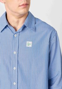 Muškarac nosi plavu i bijelu košulju s okomitim prugama i bijelim kvadratnim zakrpom na prsima na kojoj piše "LA MAISON AMV".