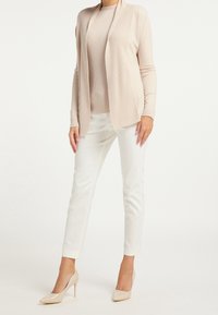 Beżowy prążkowany kardigan na jasnobeżowej dopasowanej bluzce, zestawiony z białymi obcisłymi spodniami i beżowymi szpilkami, podkreślający dopasowany wygląd.