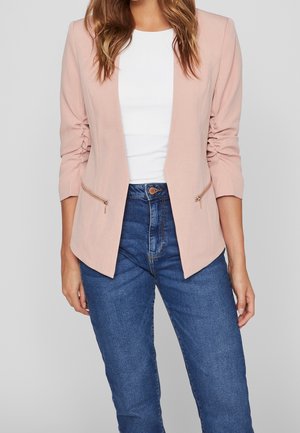 Femme portant un blazer froncé rose clair avec des poches zippées sur un top blanc et un jean bleu taille haute, debout devant un fond uni.