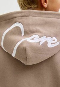 Smeđa dukserica s teksturiranom tkaninom; na stražnjoj strani kapuljače ima bijeli izvezeni logo "care".