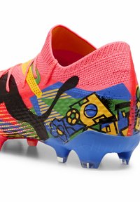 Scarpa da calcio con tomaia in maglia rosa, dettagli neri e grafiche laterali colorate. Suola testurizzata con dettagli blu e tacchetti trasparenti.