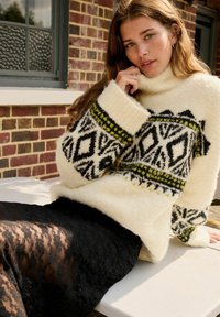 Pull ivoire et duveteux avec des motifs géométriques noirs et jaunes ; associé à une jupe en dentelle noire transparente. Détails de tissu texturé et chaud visibles.