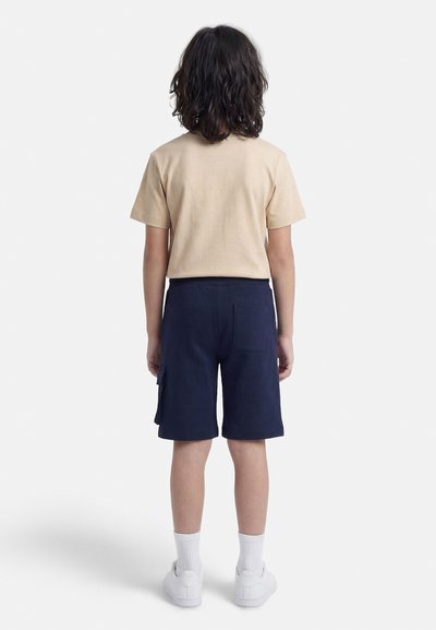 Παιδί με καστανά μαλλιά μέχρι τους ώμους στέκεται γυρισμένο πλάτη, φοράει μπεζ μπλουζάκι, ναυτικά σορτς με τσέπες, λευκές κάλτσες και λευκά αθλητικά παπούτσια.