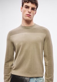 Beżowy sweter dziany z prążkowanym wzorem na górnej części ramion i rękawach, z okrągłym dekoltem i subtelnym logo na piersi.